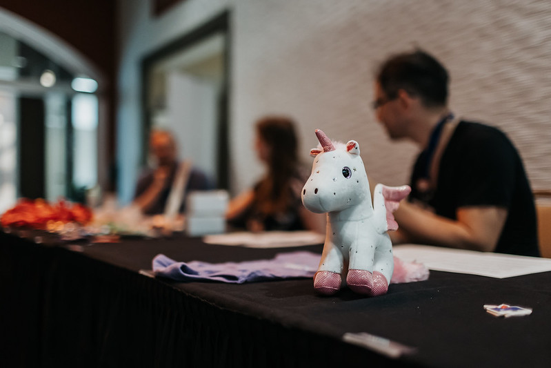 A small Django pony/unicorn on a table at DjangoCon US 2024 with blurred out organizers in the background.