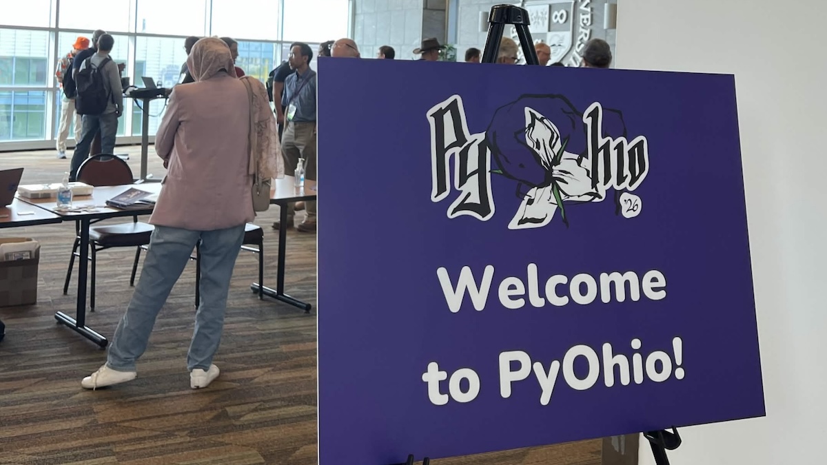A sign reading, Welcome To PyOhio with this year's logo on it. Some conference attendees are conversing in the background. Photo credit: @jonaftali@mastodon.social
